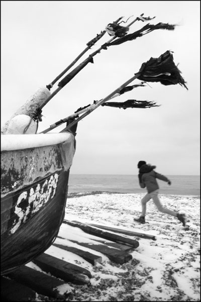 A-014. Running Against The Wind On The Snow Covered Pebbles Under The Black Flags Of Fishing Boat SM609 - by Greig Clifford